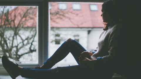 A young woman is pictured sitting on a window sill looking out: Social distancing is hard for people who already feel isolated. Photo: Pixabay