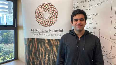 Dion Wharerau poses in front of a Te Pūnaha Matatini banner.