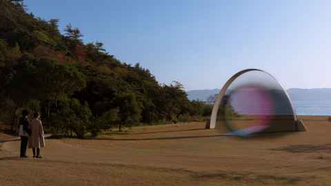 Two people stand together in a park by a lake. There is strange half-circle sculpture in the field, with a translucent rainbow orb of light inside