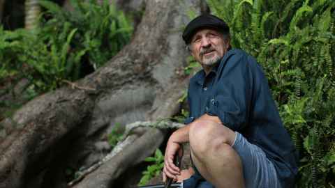 Jason Fell is wearing a cap and crouching next to a massive Moreton Bay fig tree. 