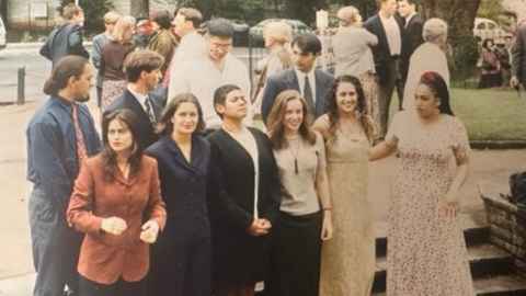 A young AP Elana Curtis is in a group shot for the MAPAS completion ceremony in 1995 outside the University of Auckland's Clocktower Building. She is second from the bottom- right row, smiling at the camera and is wearing a cream-coloured maxi dress.