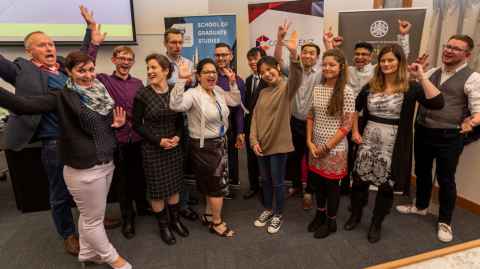 14 judges and participants wave excitedly at the camera after the 2019 Three Minute Thesis Final on 9 August.