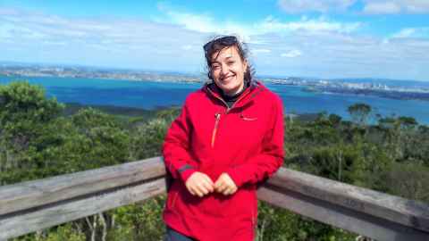 Elena, in a red coat, leans on a wooden barrier. Auckland city and the turquoise sea are in the background (taken from Rangitoto Island).
