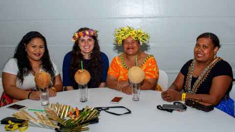 Siobhan and three other women sitting around a table, drinking from coconuts. Siobhan and another woman are wearing flower headdresses.