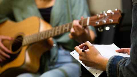 A person strums an acoustic guitar while another person writes on music manuscript paper 